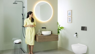 Vrouw in gele jurk voor mintgroene badkamerwand met meubel, wastafel, spiegel en wc van Geberit en zwarte kranen